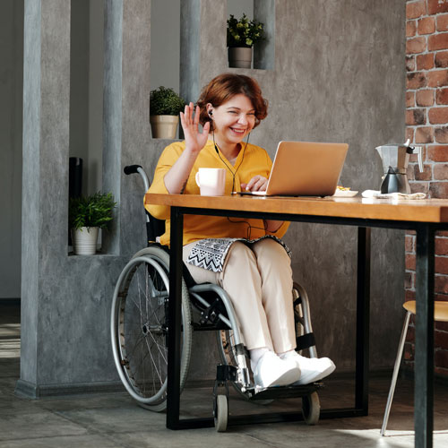A female wheelchair user sits in front of a laptop at a table in a cafe. She is smiling and waving to the screen, presumably introducing herself in a virtual meeting.